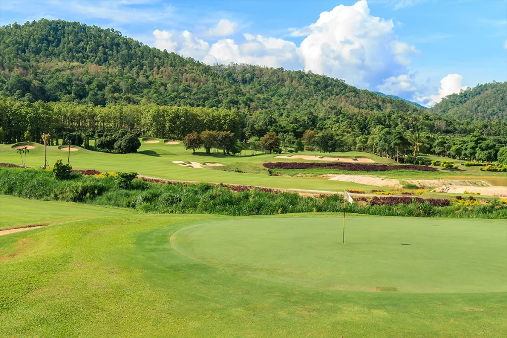 Weitläufiger Blick auf einen modernen Golfplatz mit gepflegtem Green, mehreren Sandbunkern und einer hügeligen Landschaft. Im Hintergrund ist ein dichter Wald vor blauem Himmel zu sehen.