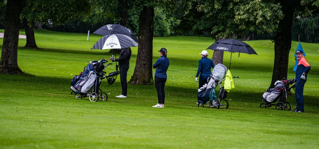 Vier Personen mit Regenschirmen warten am Fairway. Golfbags stehen auf Trolleys.