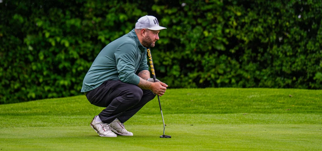 Golfer in grüner Jacke kniet auf dem Grün. Er prüft die Puttlinie und hält einen Putter.